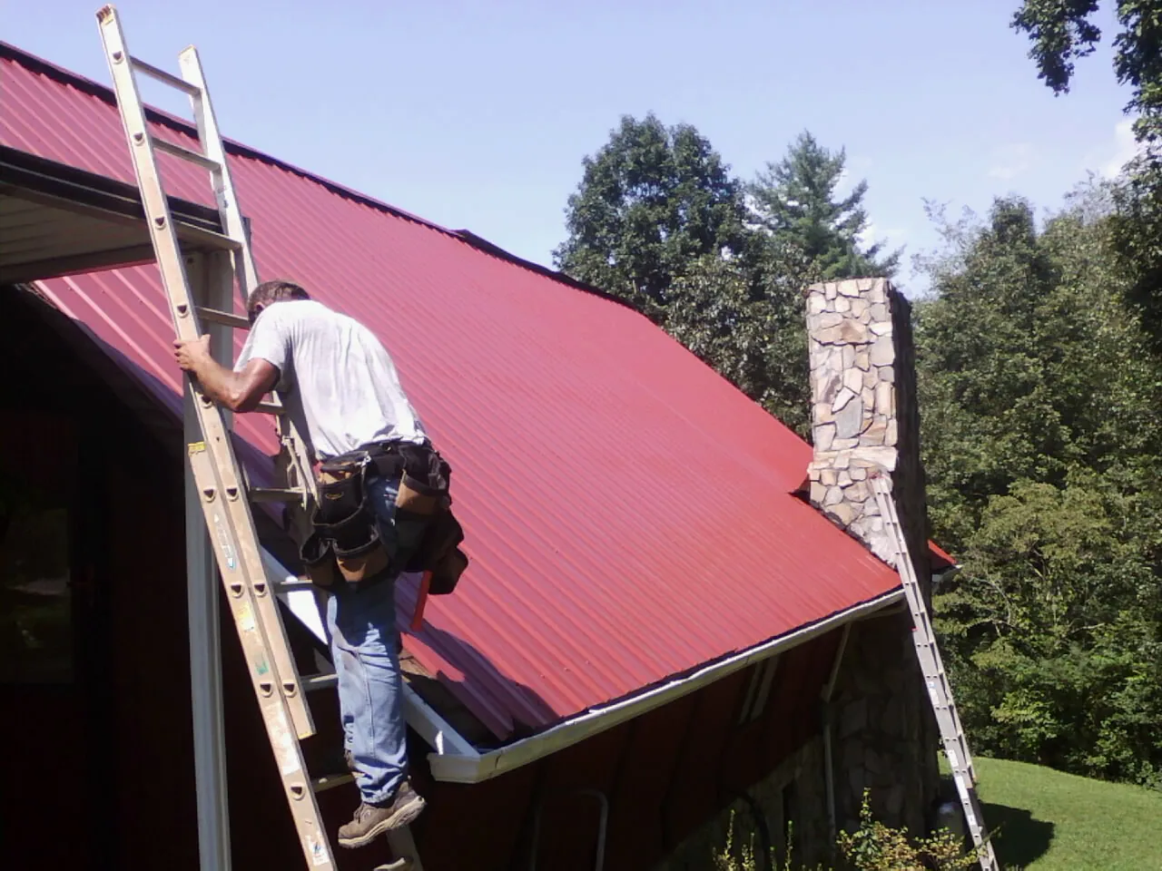 Roofer on a ladder beside a red metal roof and stone chimney — Soffit & Fascia Repair by Roofing Brothers in Mount Tabor